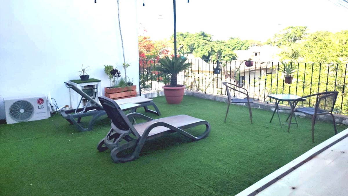 An inviting rooftop space features lounging chairs and a small table on artificial grass. Potted plants add greenery, while views of the surrounding trees are visible in the background. An air conditioning unit is installed, providing comfort for guests.