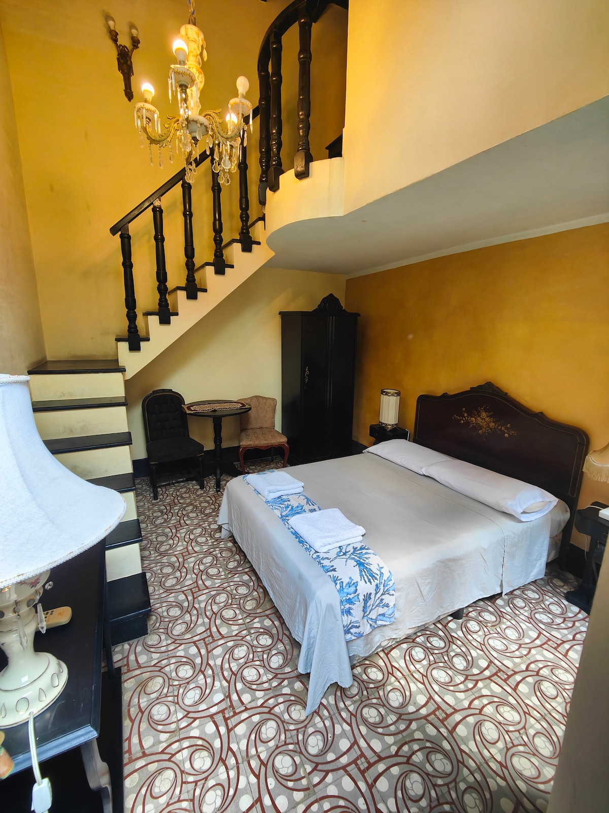 A cozy bedroom showcases a spacious bed dressed with crisp linens and accented by decorative pillows. An antique chandelier hangs from the ceiling, adding charm. A small table and chairs are positioned nearby, and a staircase leads to an upper level, enhancing the room's character.