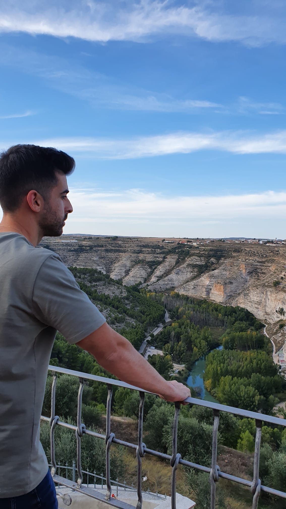 A panoramic view from a balcony showcases a vast landscape with rolling hills and lush greenery, interspersed with a river winding through the valley. The sky above is a soft blue, dotted with wispy clouds, enhancing the sense of tranquility in the scene.