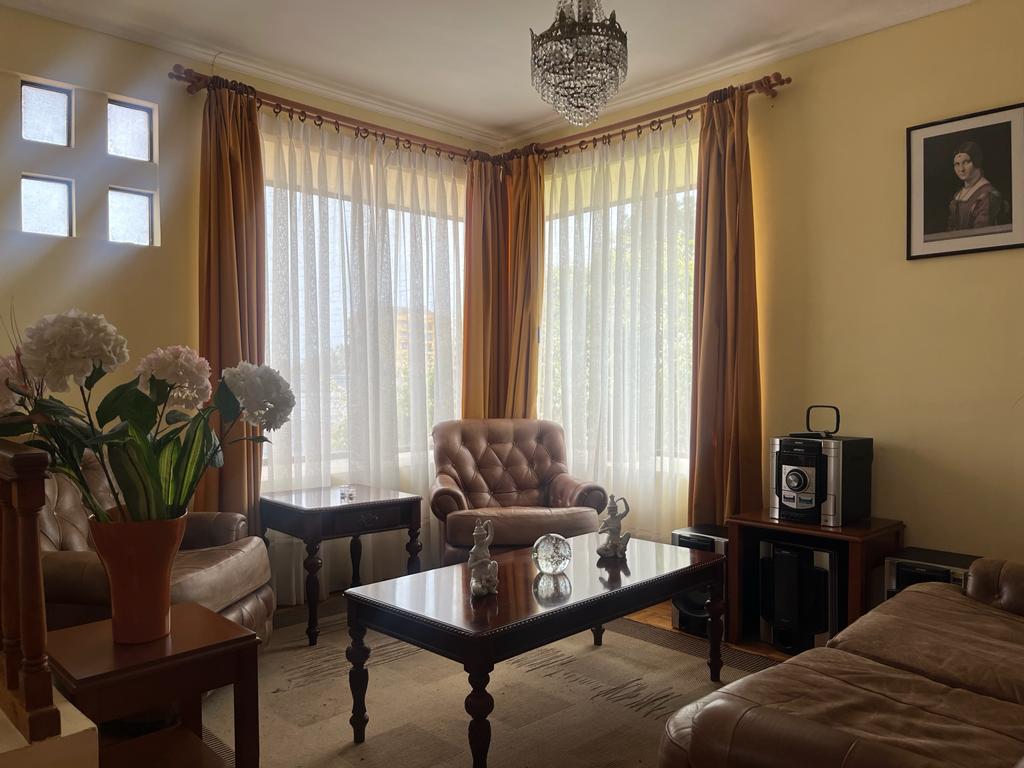 An inviting living room features large windows adorned with sheer and draped curtains, allowing natural light to fill the space. A pair of comfortable, tan armchairs sits beside a dark wooden coffee table, complemented by a decorative vase of flowers. A vintage chandelier hangs overhead.
