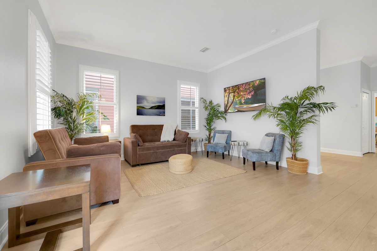 A spacious living area is furnished with a combination of brown sofas and light blue chairs, arranged around a natural fiber rug. Tall windows allow natural light to fill the room, and potted plants add a touch of greenery to the setting.