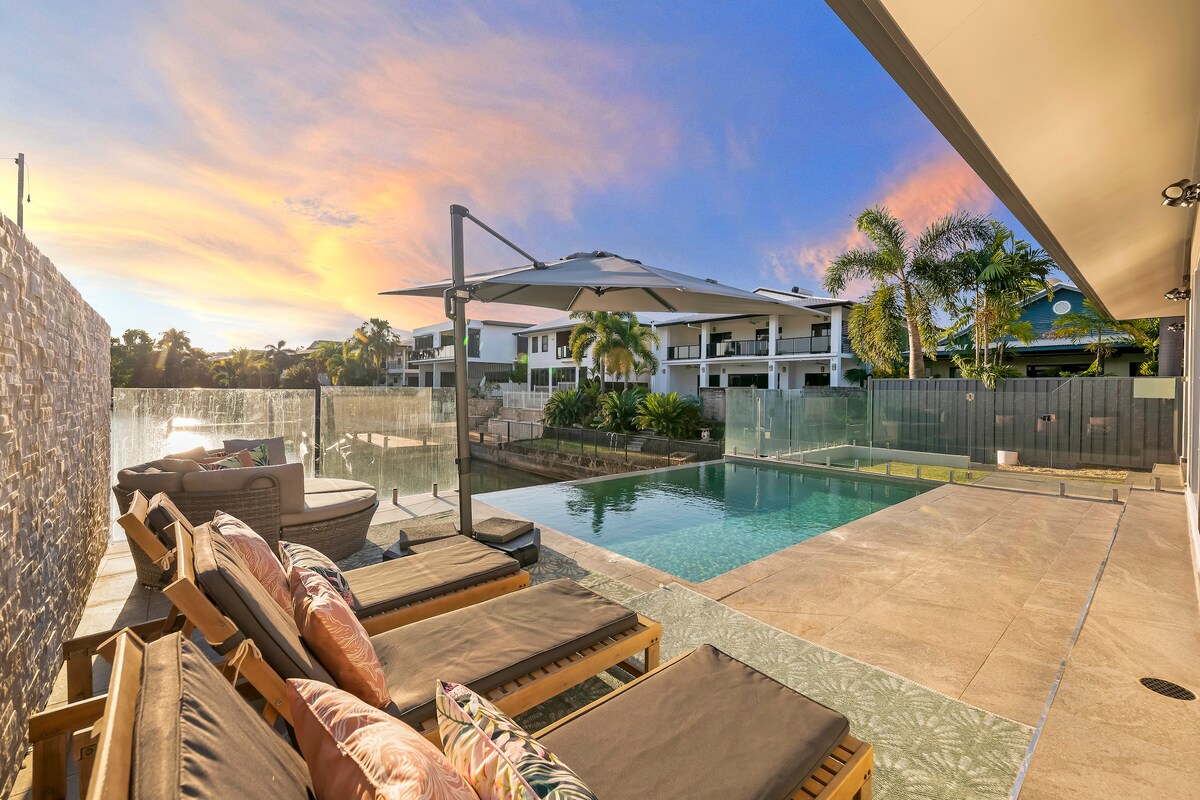 An outdoor area features comfortable lounge chairs arranged beside an infinity-edge pool, with scenic views of the marina and palm trees. The setting is enhanced by a vibrant sunset sky, creating a serene atmosphere. A large umbrella provides shade over the seating area.