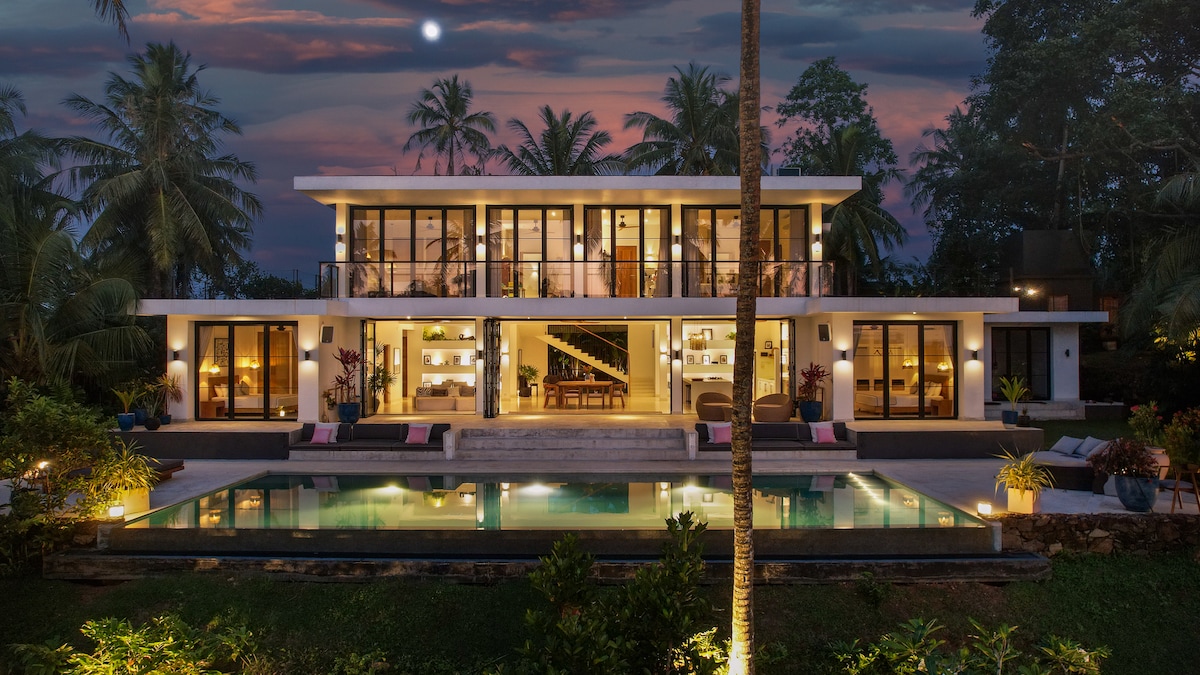The villa is presented in a modern architectural style, featuring large glass doors and expansive balconies. An inviting infinity pool reflects the evening sky, surrounded by tropical greenery. Soft lighting highlights the outdoor areas, creating a welcoming ambiance as the lush surroundings evoke a serene evening atmosphere.