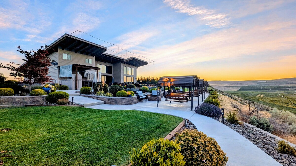 A spacious outdoor area is shown, complete with manicured lawns and various shrubs. Comfortable seating options are visible, alongside a gazebo. The backdrop features a modern building set against a vibrant sunset that casts warm colors over the Columbia River valley.