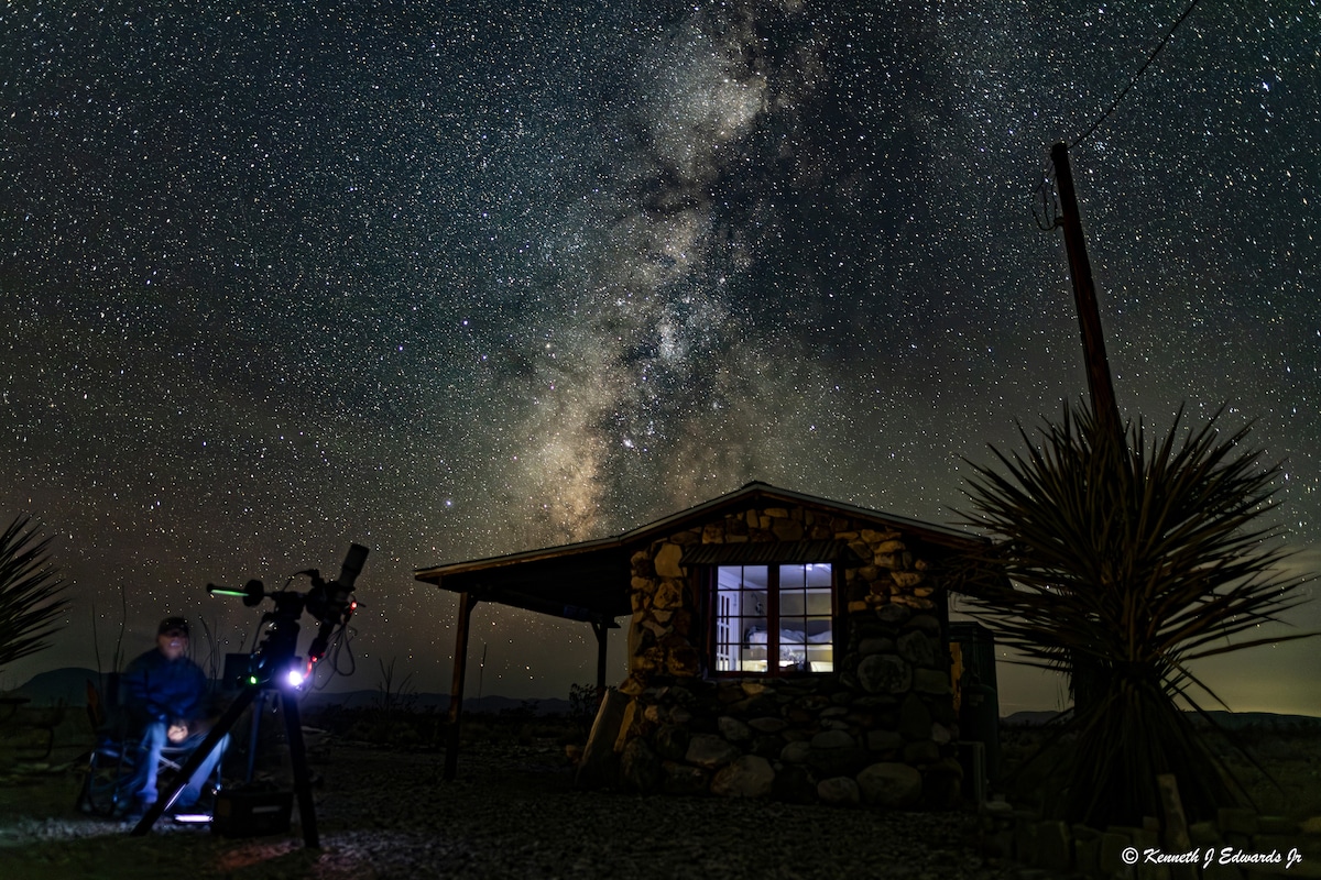 A rustic cabin is illuminated by light from its windows against a backdrop of a star-filled night sky. A person is positioned next to a telescope, with the Milky Way clearly visible above. The surrounding landscape features sparse vegetation, enhancing the secluded atmosphere.