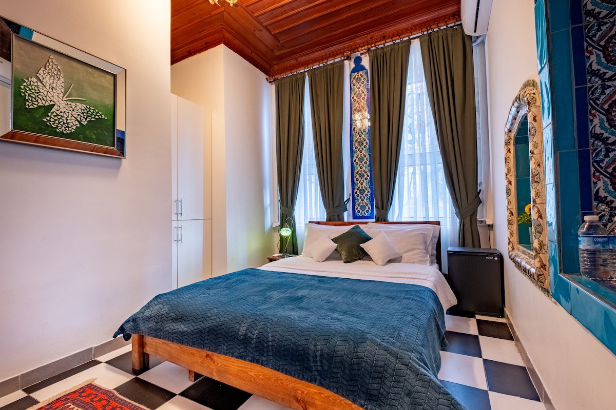 A cozy room features a spacious bed with a deep blue coverlet, complemented by decorative pillows. Tall windows draped in green curtains allow natural light to fill the space. A small refrigerator is positioned in a corner, and artistic wall decor adds character to the ambiance.