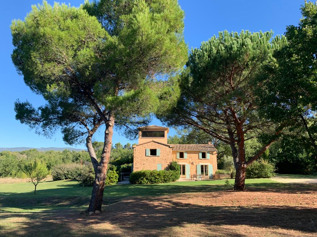 A charming Provençal house is nestled among tall trees in a spacious green park. A clear blue sky sets the backdrop, highlighting the rustic stone exterior and large windows of the home, which is surrounded by well-maintained grounds and natural landscaping.