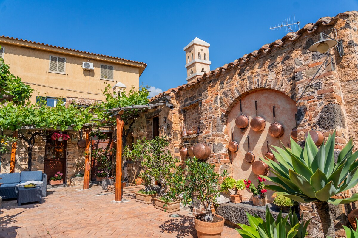 A charming outdoor area is surrounded by stone walls, featuring a variety of plants and decorative copper pots. Comfortable seating is arranged on terracotta tiles, and a trellis covered with greenery adds to the pleasant ambiance. A view of a bell tower is visible in the distance.