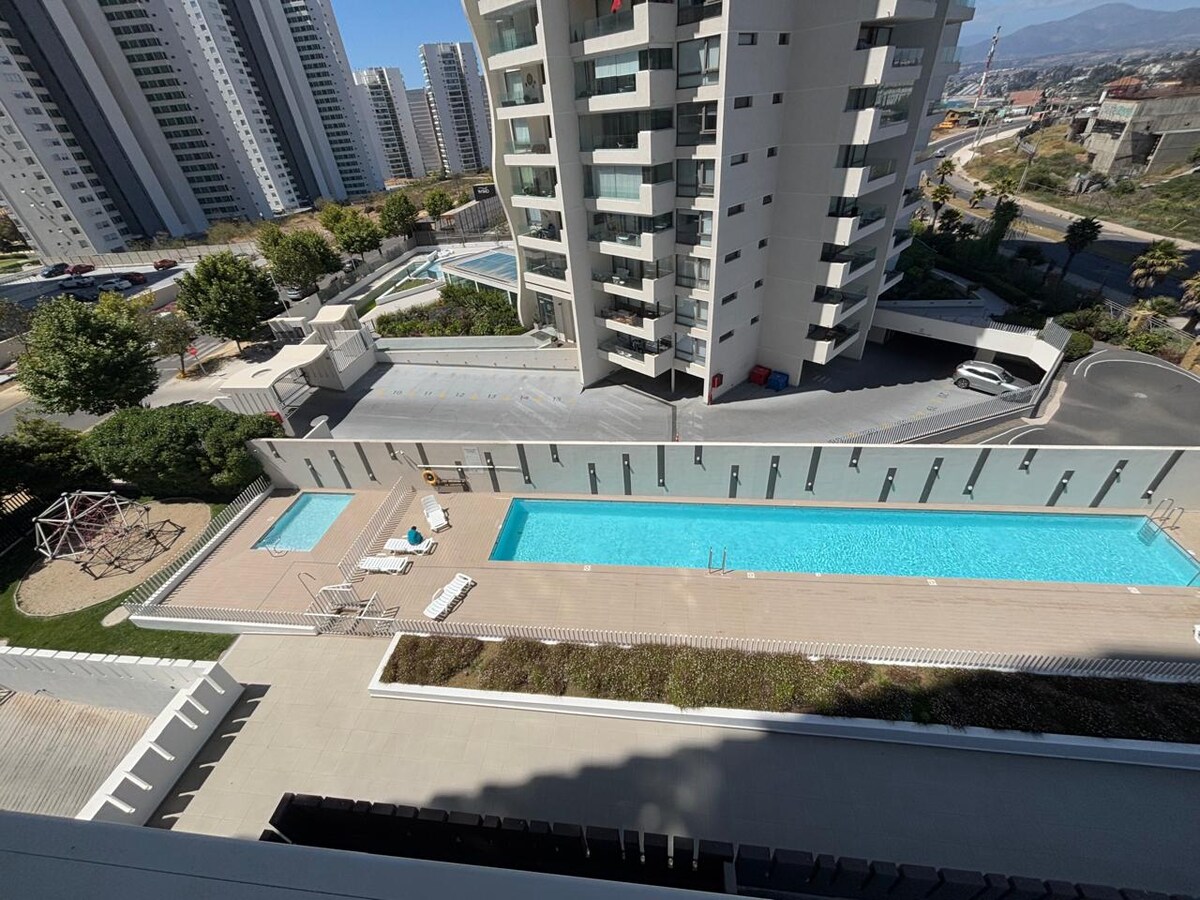 An aerial view showcases the outdoor semi-olympic pool, surrounded by a clean patio area with lounge chairs. Nearby, a playground is visible among the greenery, while modern buildings outline the background against a clear sky.