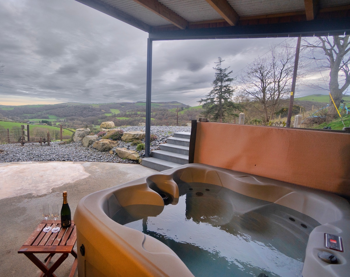 A modern hot tub is positioned on a patio, overlooking rolling green hills under a cloudy sky. A small wooden table nearby is set with a bottle of champagne and glasses, providing an inviting space for relaxation amidst the serene landscape.