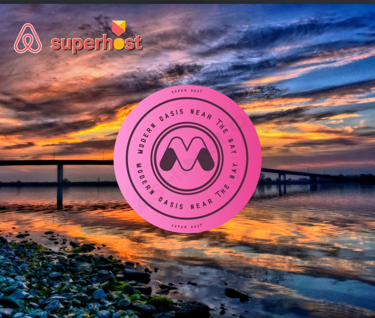 A vibrant sunset reflects on calm waters near a shoreline, with a bridge in the background. The sky showcases a blend of warm colors, including oranges and purples, enhancing the serene atmosphere of the bay.