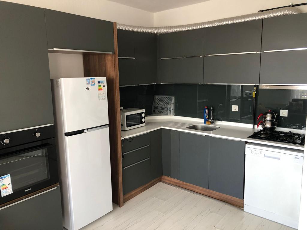 The modern kitchen space features sleek, grey cabinetry and a large window providing natural light. Stainless steel appliances, including a refrigerator, dishwasher, and oven, are included. A microwave and sink are also present, creating a fully equipped cooking area.