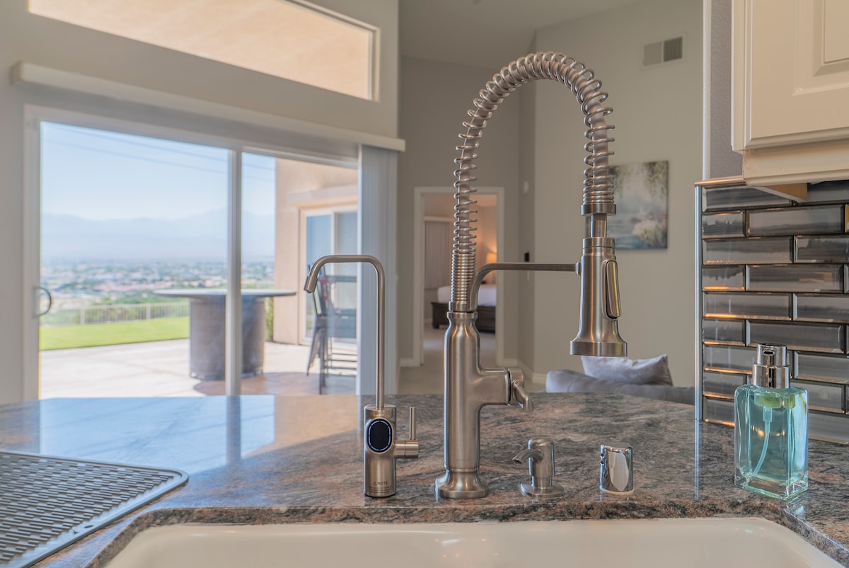 A modern kitchen sink area is highlighted, showcasing a sleek, metallic faucet with a pull-down feature. The background reveals expansive glass doors opening to a breathtaking view of the landscape beyond, allowing natural light to enhance the space.