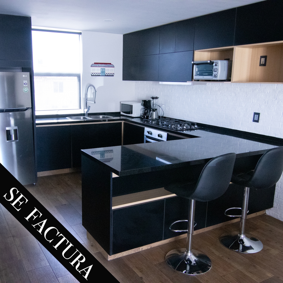 A modern kitchen is equipped with sleek black cabinetry and stunning granite countertops. It features stainless steel appliances, including a refrigerator, stove, and microwave. A small breakfast bar with two high chairs is visible, enhancing the functional layout. Natural light enters through a nearby window.