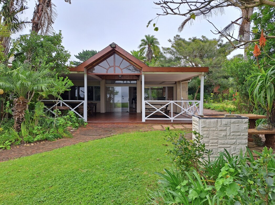 A spacious bungalow features a welcoming entrance with a high-pitched roof and large glass windows, allowing for natural light. Lush greenery surrounds the home, complemented by a manicured lawn and a stone picnic table nearby.