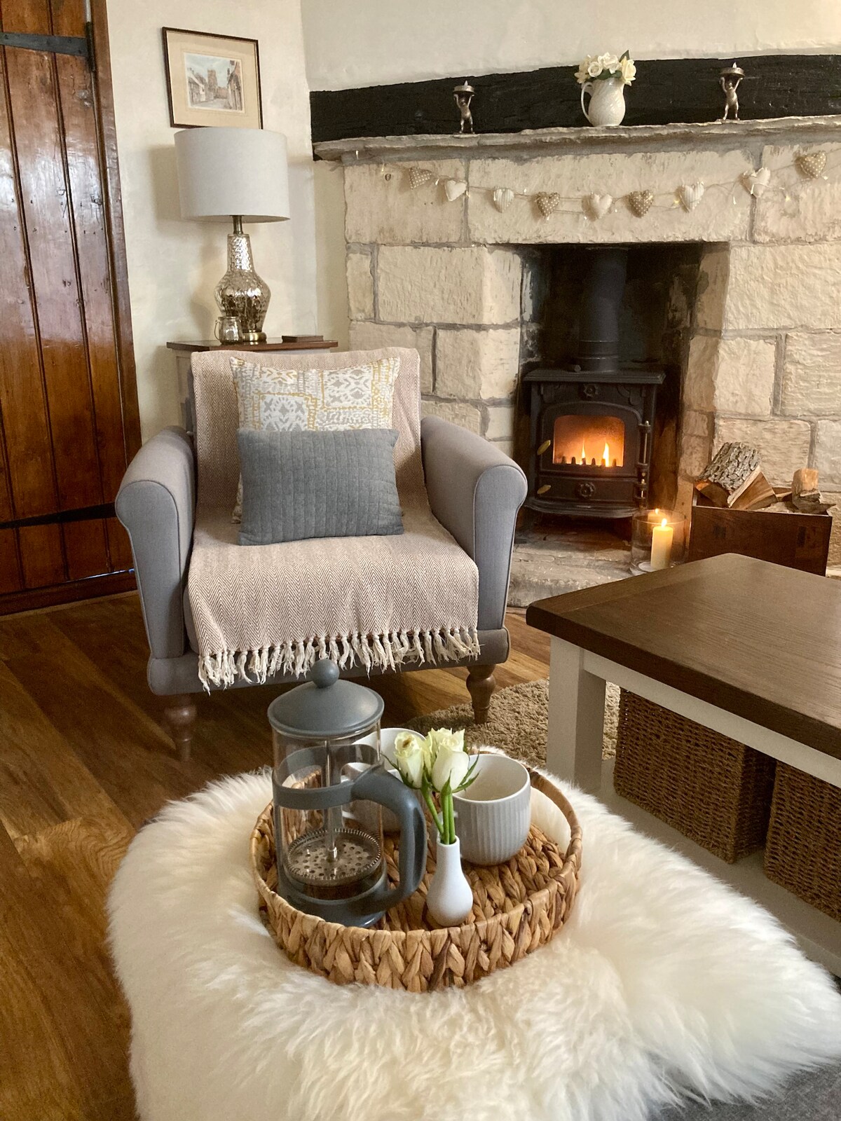 A comfortable armchair is positioned near a decorative stone fireplace, where a gentle glow of a fire can be seen. A cozy throw blanket and decorative pillows add to the chair's appeal. A rustic coffee table is adorned with a woven tray containing a lantern and fresh flowers.