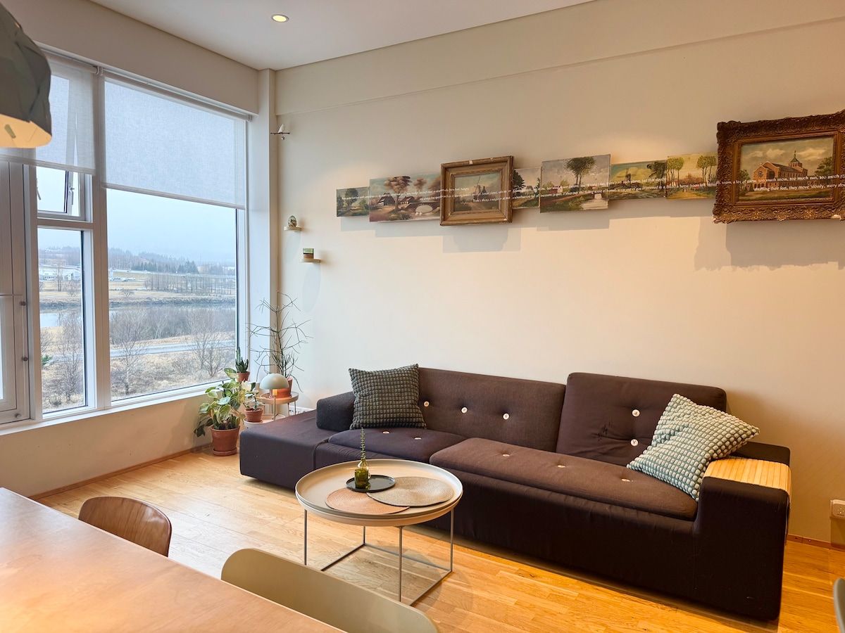 A modern living area features a dark sectional sofa adorned with patterned cushions. A round coffee table sits in front, and artwork is displayed along the wall. Natural light filters in through large windows, providing a view of the landscape outside.