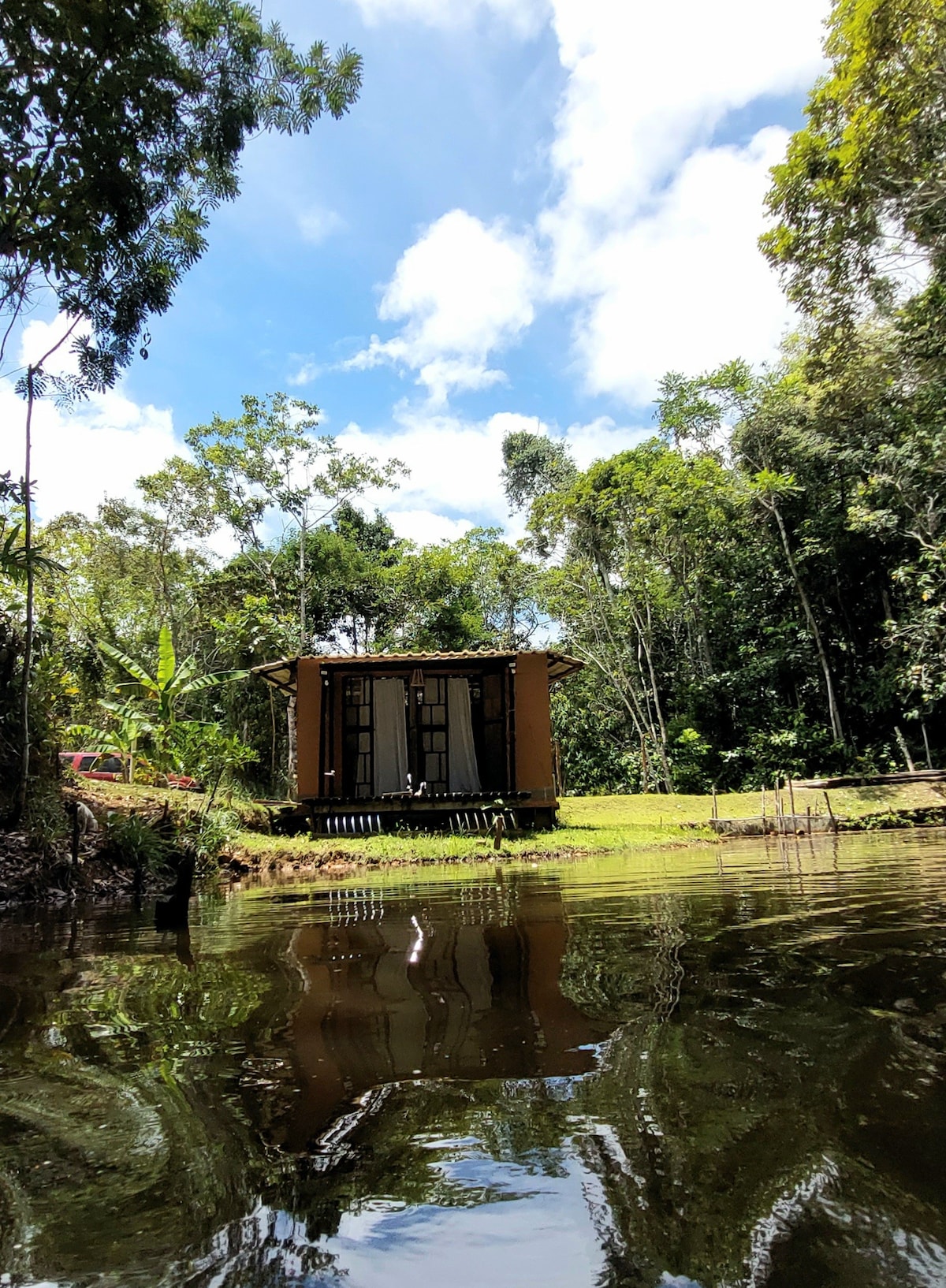 A charming chalet is viewed from the lake, surrounded by lush greenery. The structure features double doors opening onto a small deck. Reflections of the trees complement the serene water, creating a peaceful atmosphere within the natural setting.