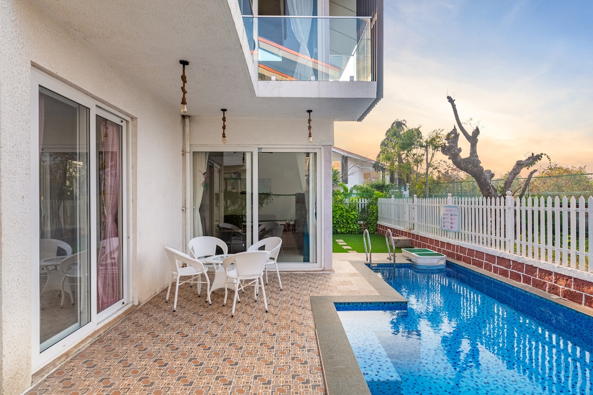 An outdoor area is highlighted, featuring a private swimming pool with a jacuzzi beside it. A patio with white chairs and a table is situated next to large glass doors that provide access to the villa's interior. Lush greenery can be seen in the background.