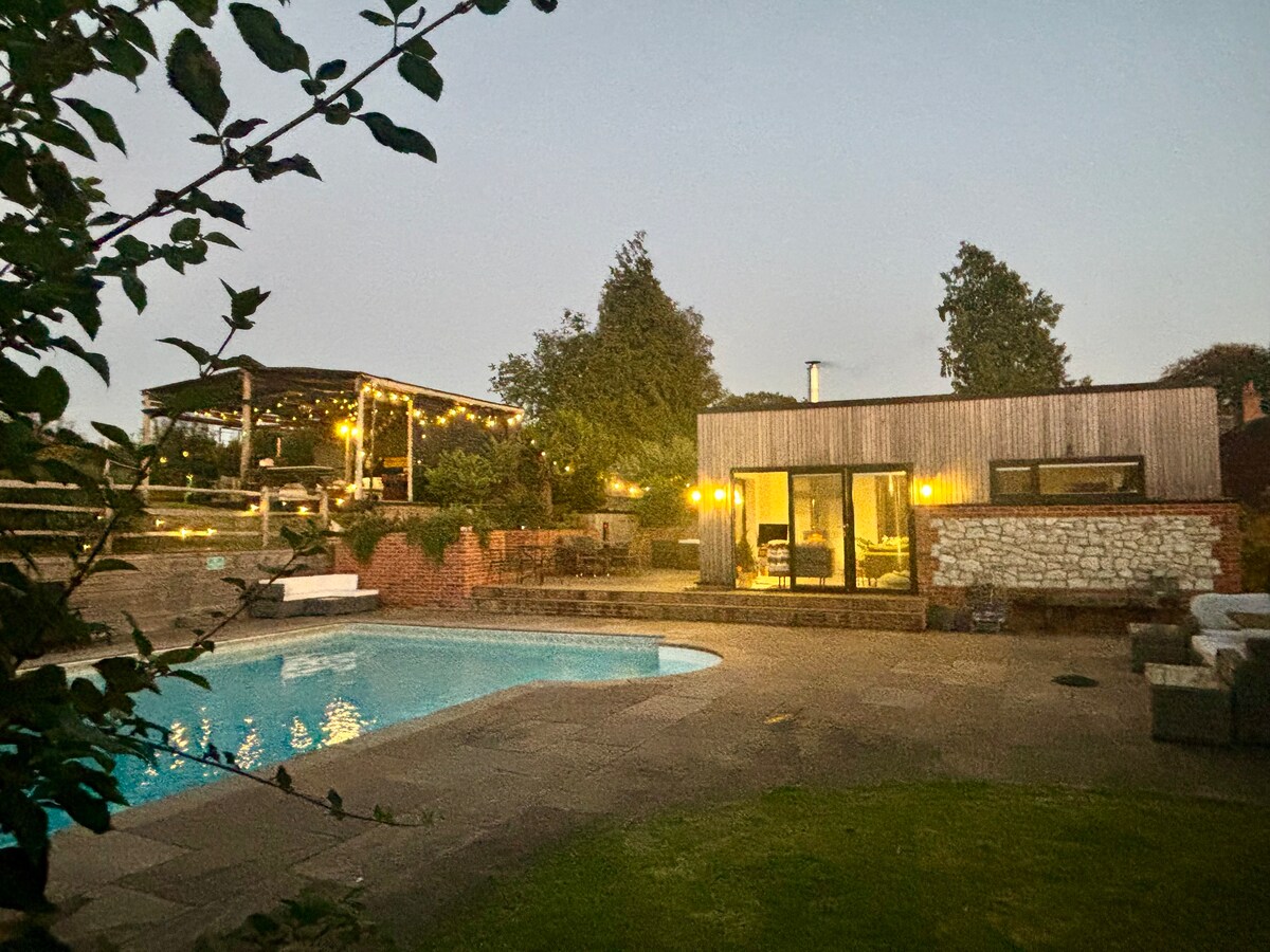 The modern pool house is illuminated during twilight, featuring large glass doors that open to a private pool. The surrounding area includes a spacious patio and an outdoor seating space, creating a peaceful setting. Lush greenery frames the scene, enhancing the inviting atmosphere.