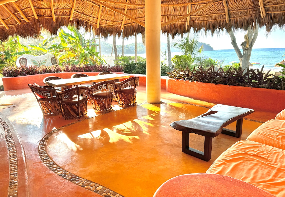 A vibrant outdoor seating area features a thatched roof overhead, surrounded by lush greenery. A spacious table stands at the center, accompanied by intricately designed chairs. The warm orange floor reflects sunlight, creating a welcoming ambiance with views of the ocean in the background.