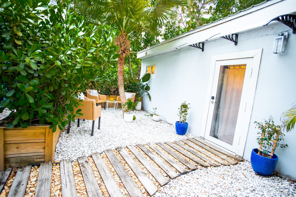 A pathway of wooden planks leads to a glass door of the guesthouse, surrounded by landscaped gravel and lush greenery. Potted plants and a small seating area with wicker chairs create a serene outdoor space, inviting relaxation in a tranquil setting.