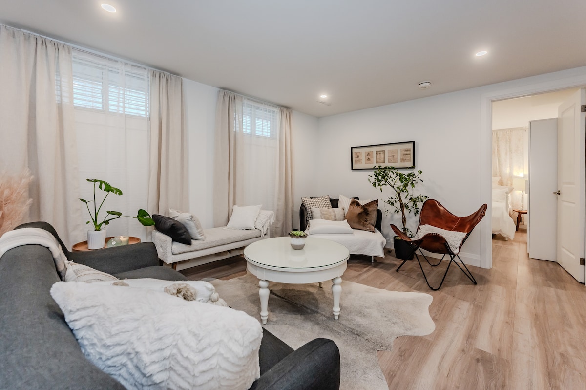 The living room features a light and airy atmosphere, with natural light filtering through sheer curtains. A round coffee table is centered among cozy seating, including a plush sofa and a stylish chair. Decorative plants and soft textiles add warmth to the space.