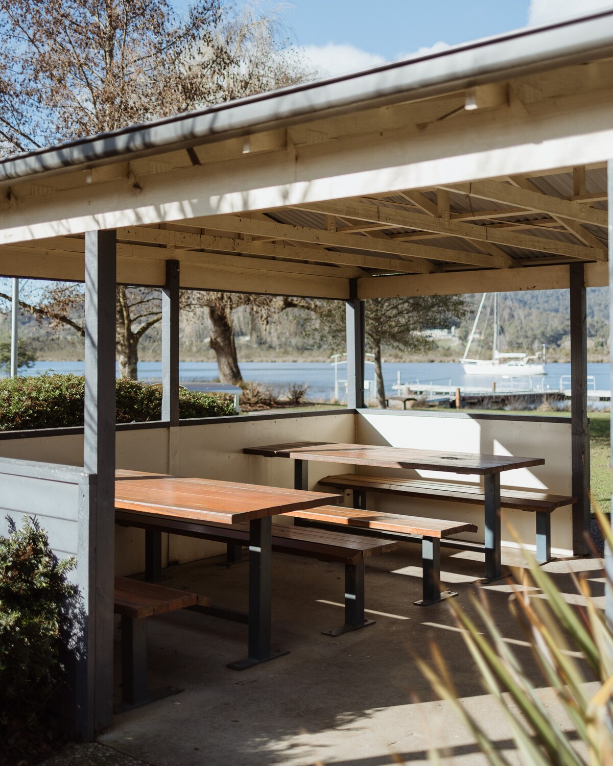 A covered outdoor seating area is present, featuring wooden tables and benches, designed for communal dining. The space overlooks the river, providing a scenic view of the water and surrounding nature.