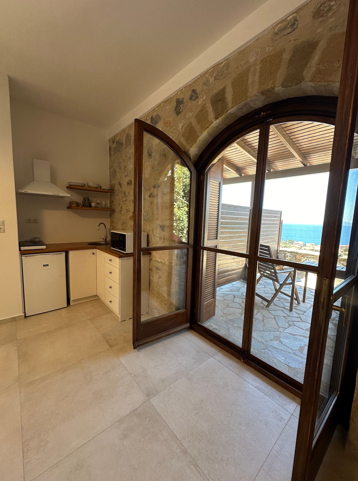 A bright kitchen area features natural stone walls and wooden cabinetry. Large glass doors open to a scenic view, allowing natural light to enter. A small refrigerator and a microwave are present, with a compact dining space visible beyond the doors.
