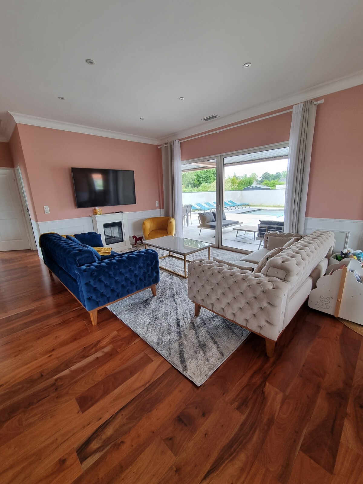 A spacious living room features plush seating in blue and beige, arranged around a glass coffee table. Large windows allow natural light to fill the space, revealing a view of the outdoor terrace and pool area. A television is mounted on the wall opposite the seating.