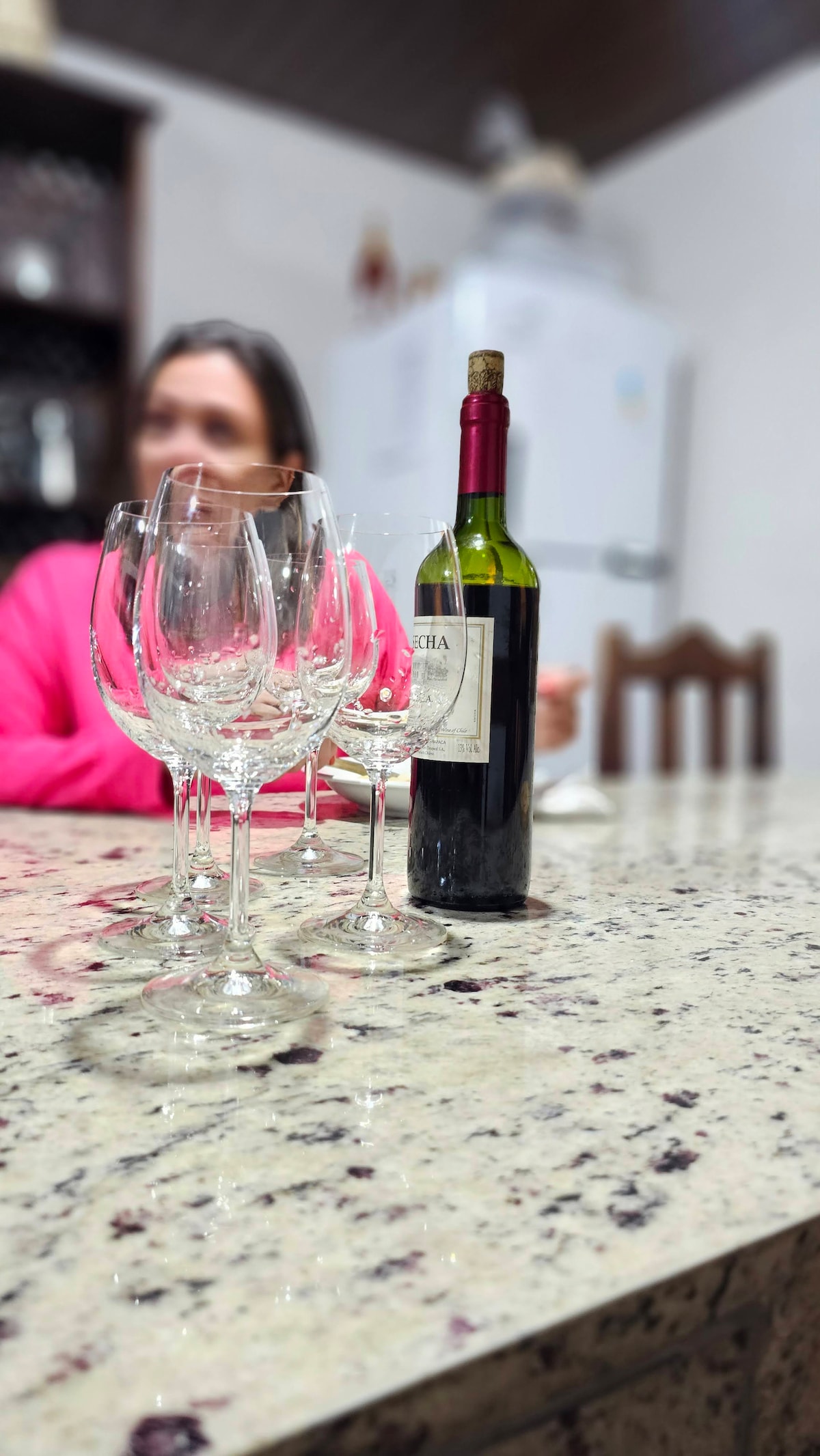 A granite countertop holds a bottle of wine alongside several clear wine glasses. The background features a white refrigerator and wooden dining chairs, creating a warm and inviting setting for gatherings.