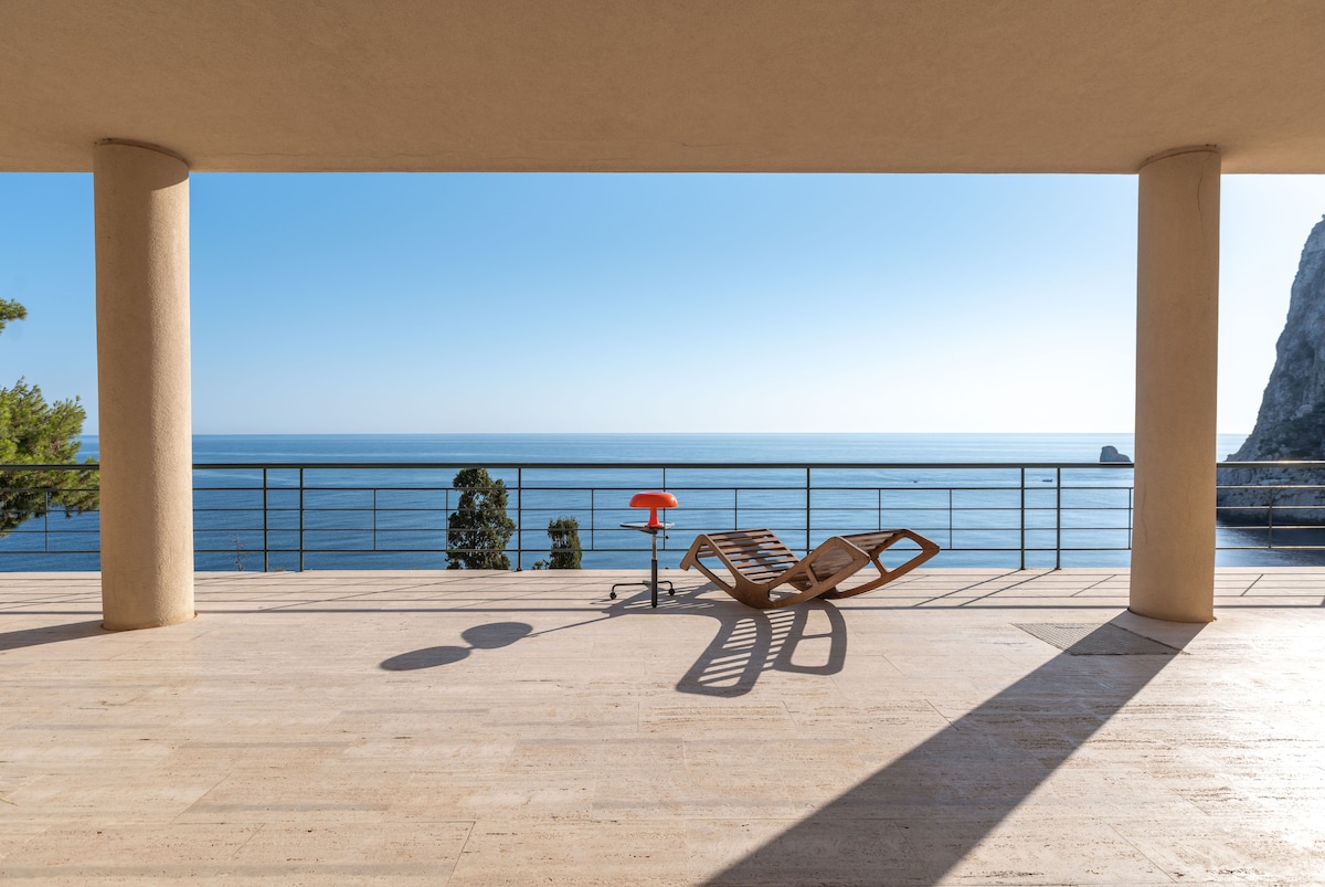 A spacious terrace offers unobstructed views of the open sea and distant cliffs. A modern lounge chair and a small side table are positioned on the light-colored stone flooring. Sunlight casts soft shadows, enhancing the serene atmosphere of the outdoor space.