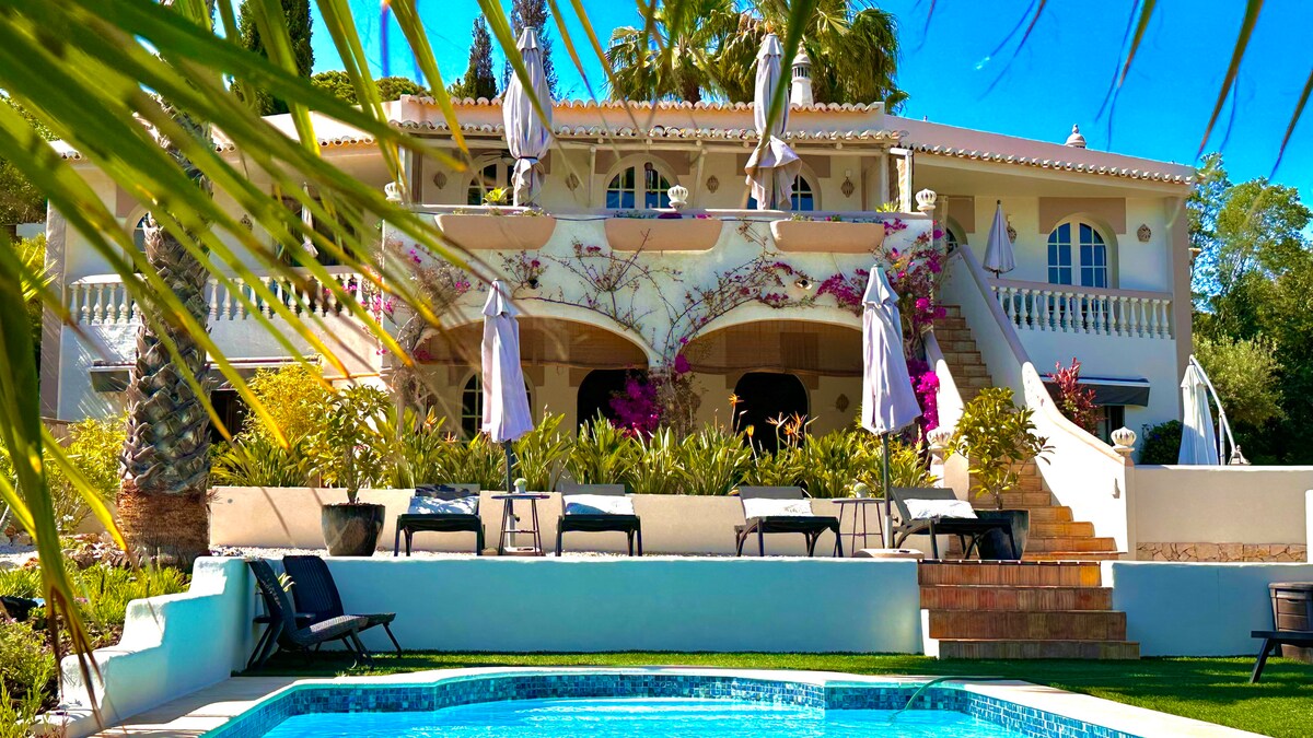 The villa's elegant exterior is framed by lush palm trees and vibrant floral arrangements. A refreshing pool is visible in the foreground, with lounge chairs arranged for relaxation. The expansive terrace above features several umbrellas, providing shade and enhancing the outdoor seating area.