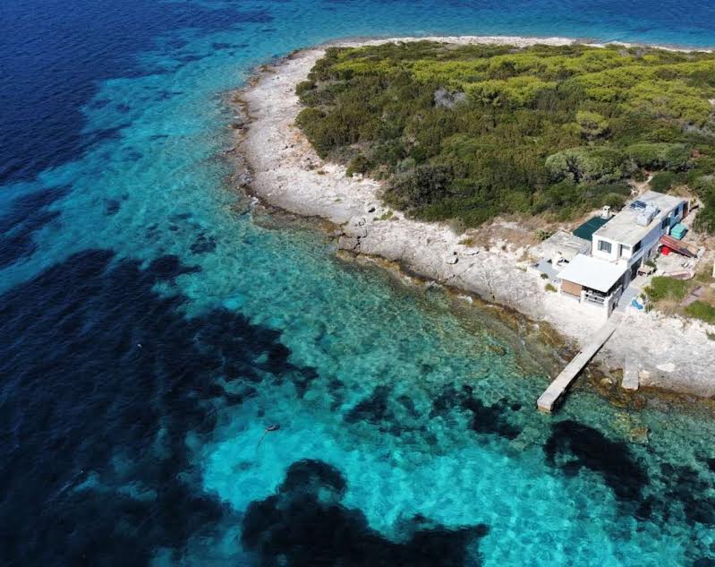 An aerial view showcases the island's rugged shoreline, where clear turquoise waters meet rocky edges. A small dock extends into the ocean, leading to a modest house surrounded by lush greenery. The pristine landscape emphasizes the island's natural beauty and tranquility.