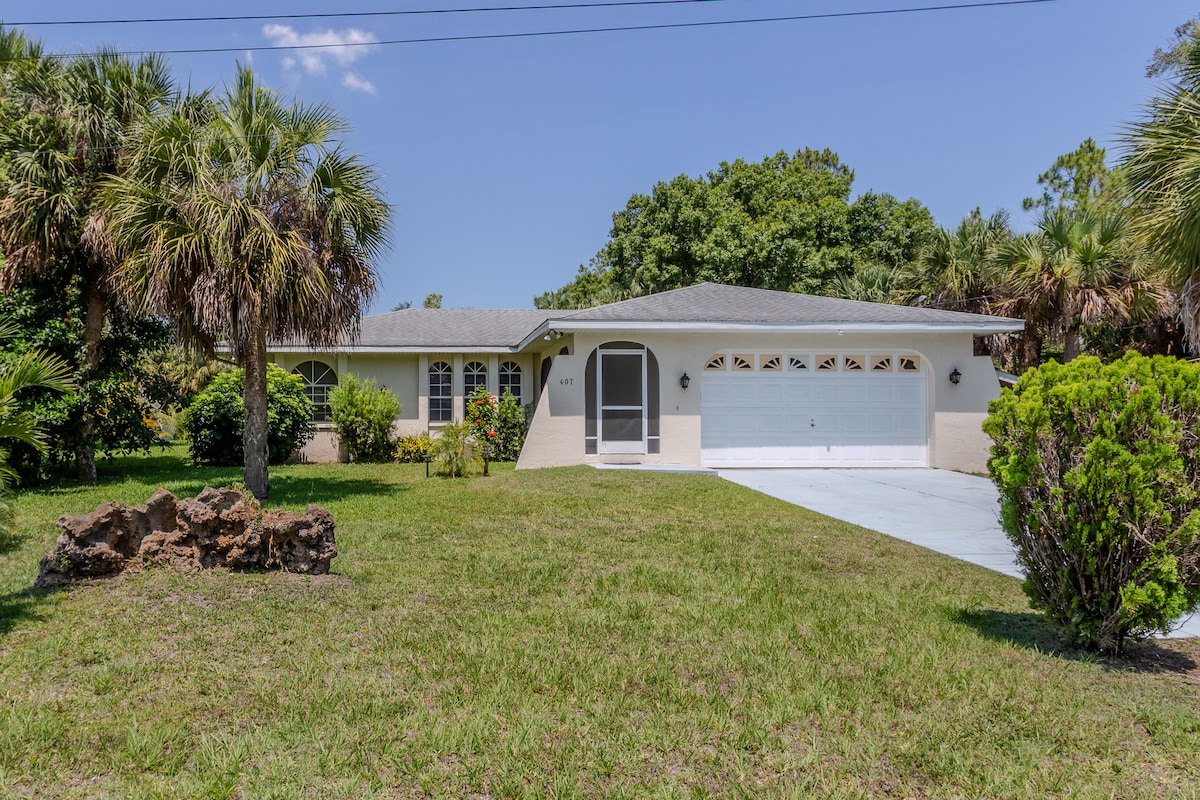 The exterior of the home is showcased, featuring a well-maintained front yard with lush greenery and palm trees. A driveway leads to a two-car garage, and a covered entryway provides access to the house. The roof is light gray, complementing the beige facade.