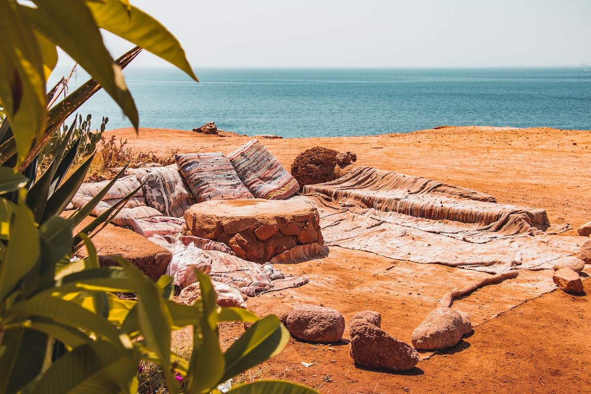 A relaxing outdoor seating area is depicted with stone structures and woven textiles arranged over the ground. Natural vegetation frames the scene, providing a connection to the serene ocean view in the background, with calm waters extending towards the horizon.