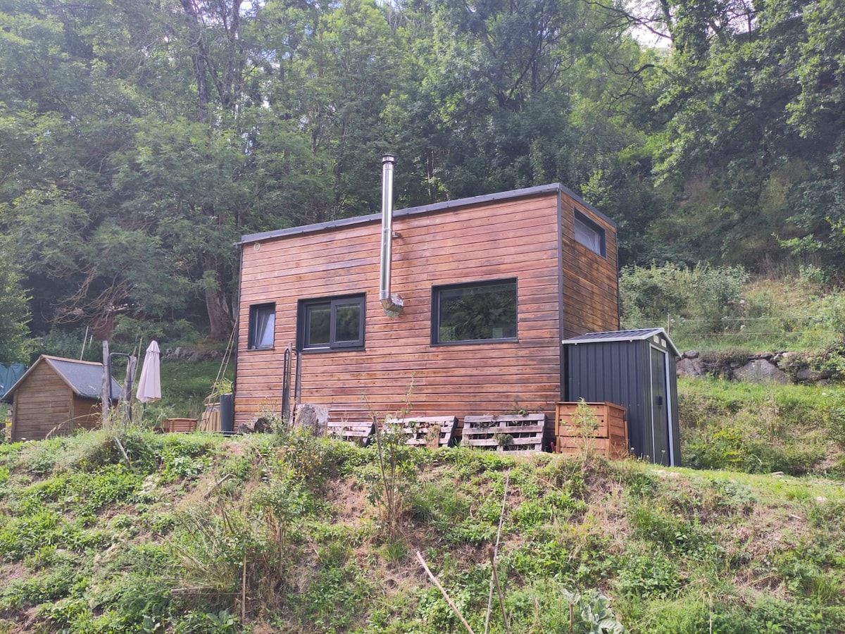 The tiny house is nestled among trees, featuring a wooden exterior with large windows. A chimney is visible on the side, indicating a wood stove inside. Outdoor seating made from pallets is arranged in front, providing a rustic space to enjoy the natural surroundings.