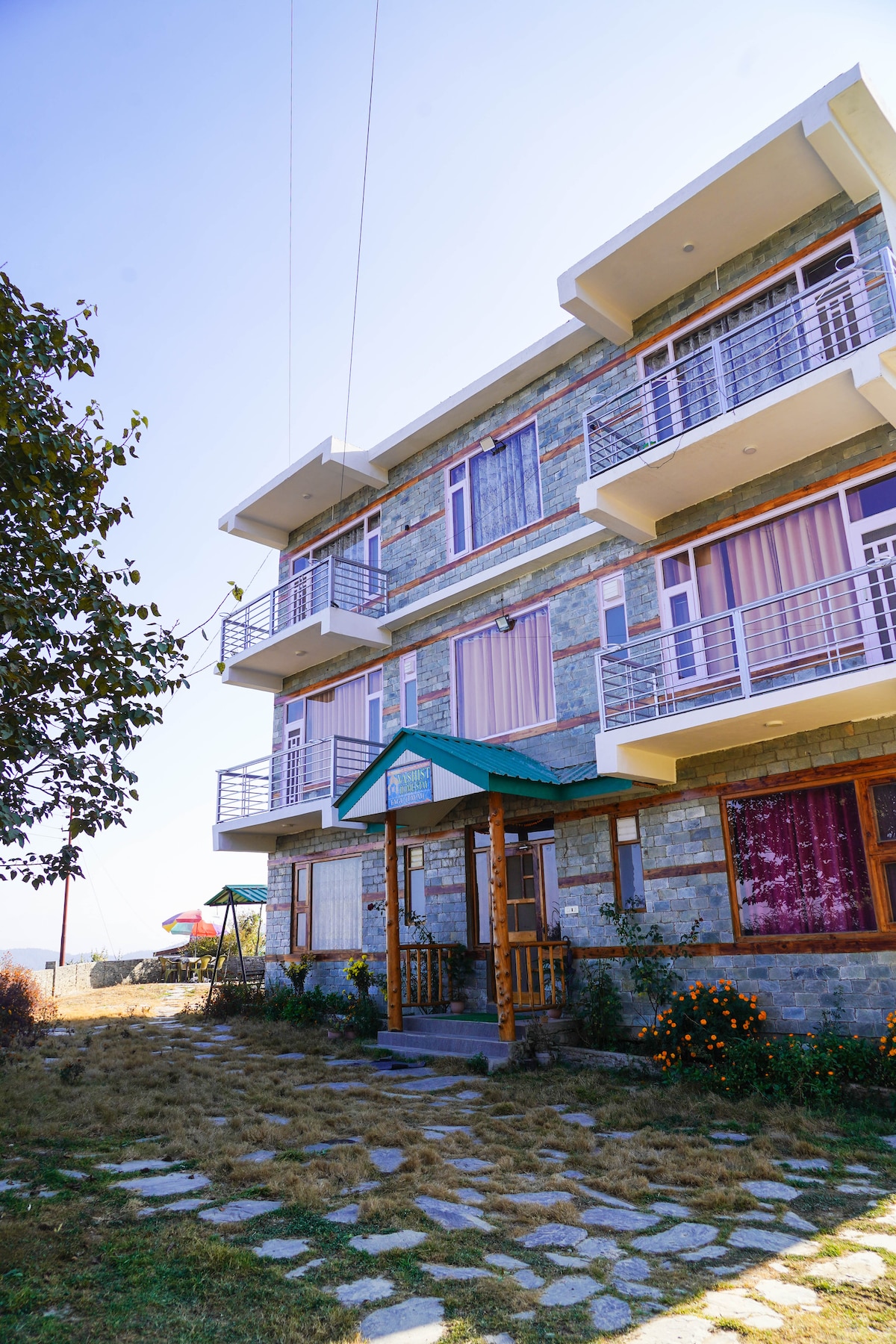 The exterior of the three-story building features a stone facade with multiple balconies, each adorned with colorful curtains. A welcoming entrance is framed by wooden elements, while a landscaped area with pathways and greenery enhances the natural setting.
