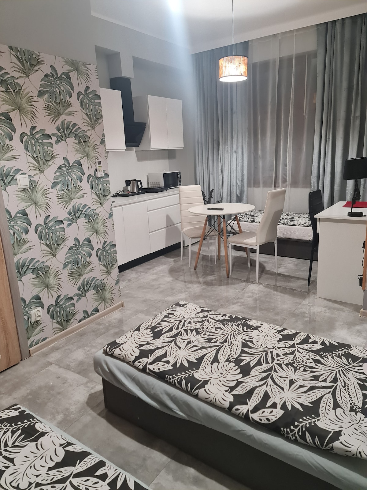 A functional room features three single beds with floral bedding. A kitchenette with modern appliances and a dining table for four is visible. Seating arrangements include black and white chairs. Light filters through sheer curtains, highlighting the green patterned wall.