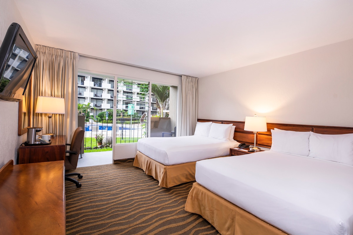 A well-lit room features two spacious beds with crisp white linens, complemented by wooden headboards. A desk and chair are positioned against a wall, while large glass doors open to a private balcony overlooking a pool area surrounded by greenery.