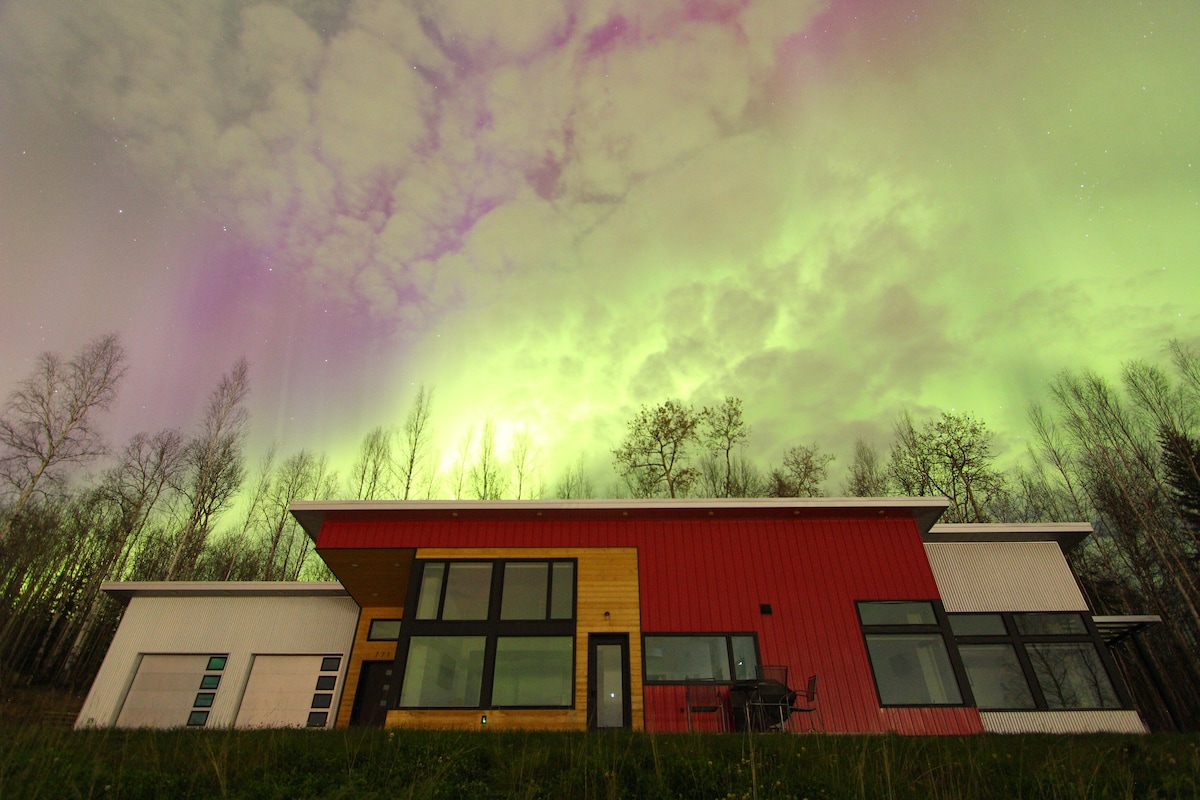 A modern home is viewed from a distance against the backdrop of the night sky illuminated by the Northern Lights. The house features large windows and a striking red and white exterior, surrounded by lightly forested terrain.
