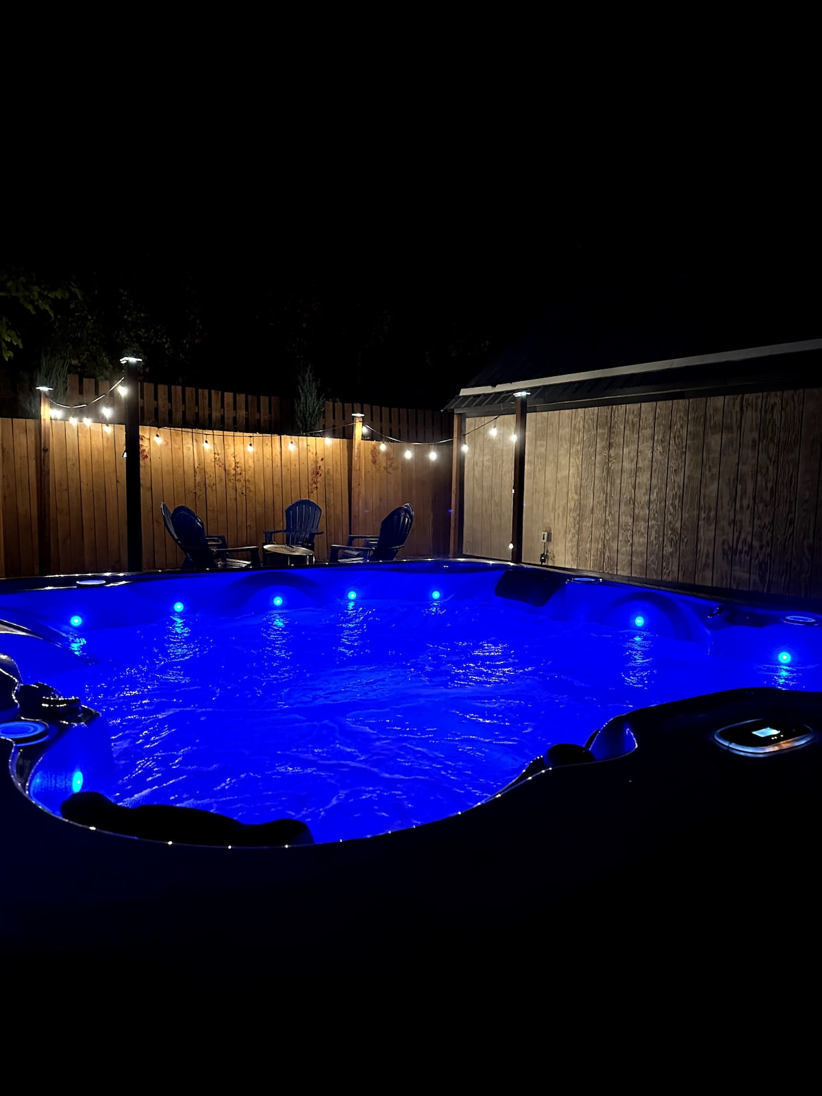 The hot tub is illuminated in a calming blue glow, surrounded by a wooden fence and string lights that add a soft ambiance. Comfortable seating is visible in the background, inviting relaxation under the night sky.