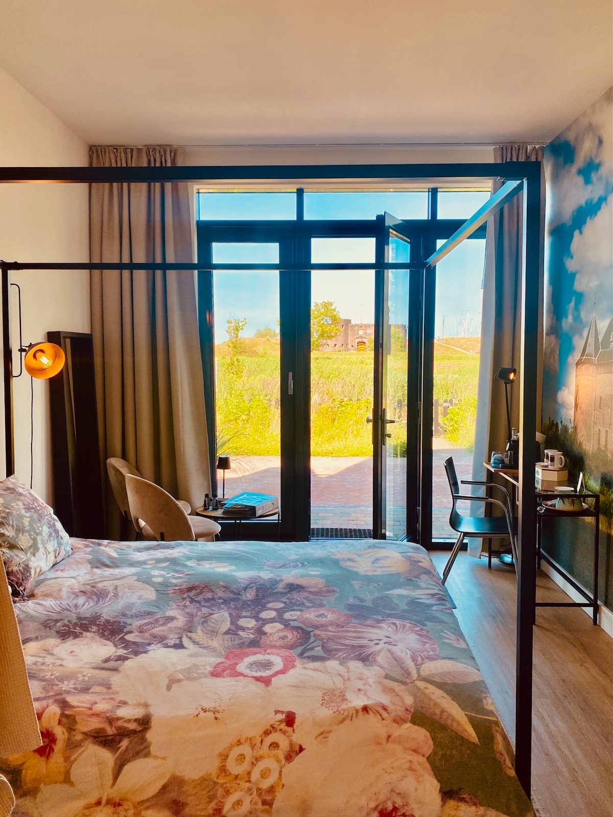 A spacious bedroom features a large floral-patterned bedspread and a black metal bed frame. Floor-to-ceiling glass doors open to a patio, allowing natural light and garden views. A small desk and chair are situated nearby, with a cozy armchair in the corner.