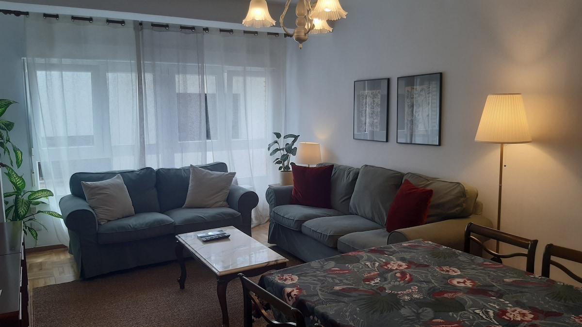 A comfortable living area features two sofas with cushions in a blend of muted colors. A coffee table sits at the center, while a dining table is visible nearby. Soft light is provided by a standing lamp and a ceiling fixture, and sheer curtains allow natural light to filter through.