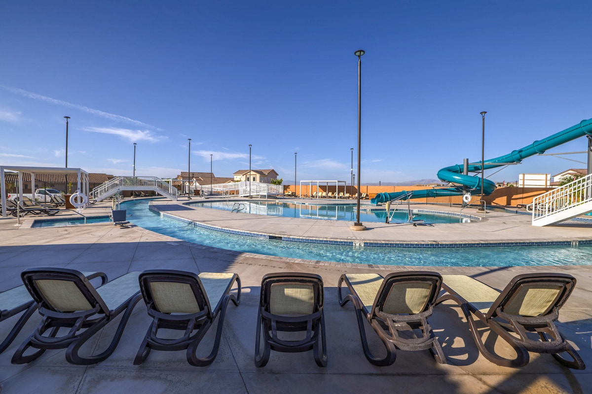 An outdoor shared waterpark features a large swimming pool with a waterslide and lazy river. Several lounge chairs line the poolside, and bright blue skies are visible above. Pathways are designed for easy access to various pool amenities.