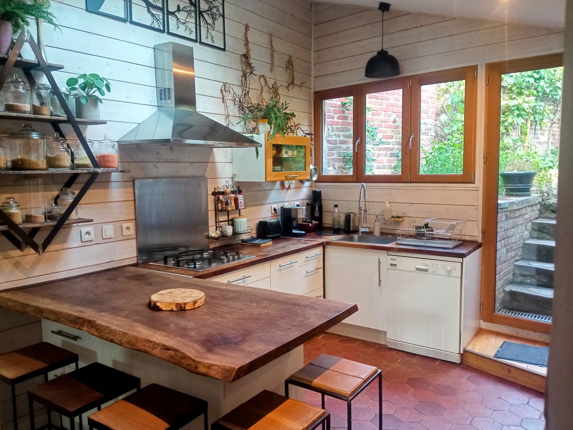 The kitchen features a wooden countertop and modern appliances, including a dishwasher and a stove. Large windows provide natural light and a view of the garden. Open shelves display various kitchenware, while a small dining area is arranged with several stools.
