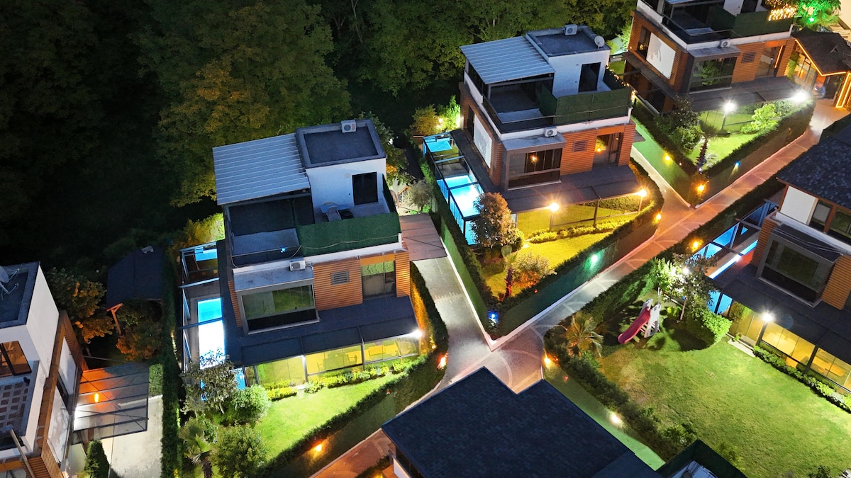 The aerial view showcases a collection of modern, independent villas surrounded by lush greenery. Each villa features a private pool, with gentle lighting illuminating the pathways. The serene atmosphere suggests a peaceful retreat, emphasizing the well-maintained landscaping and open space.