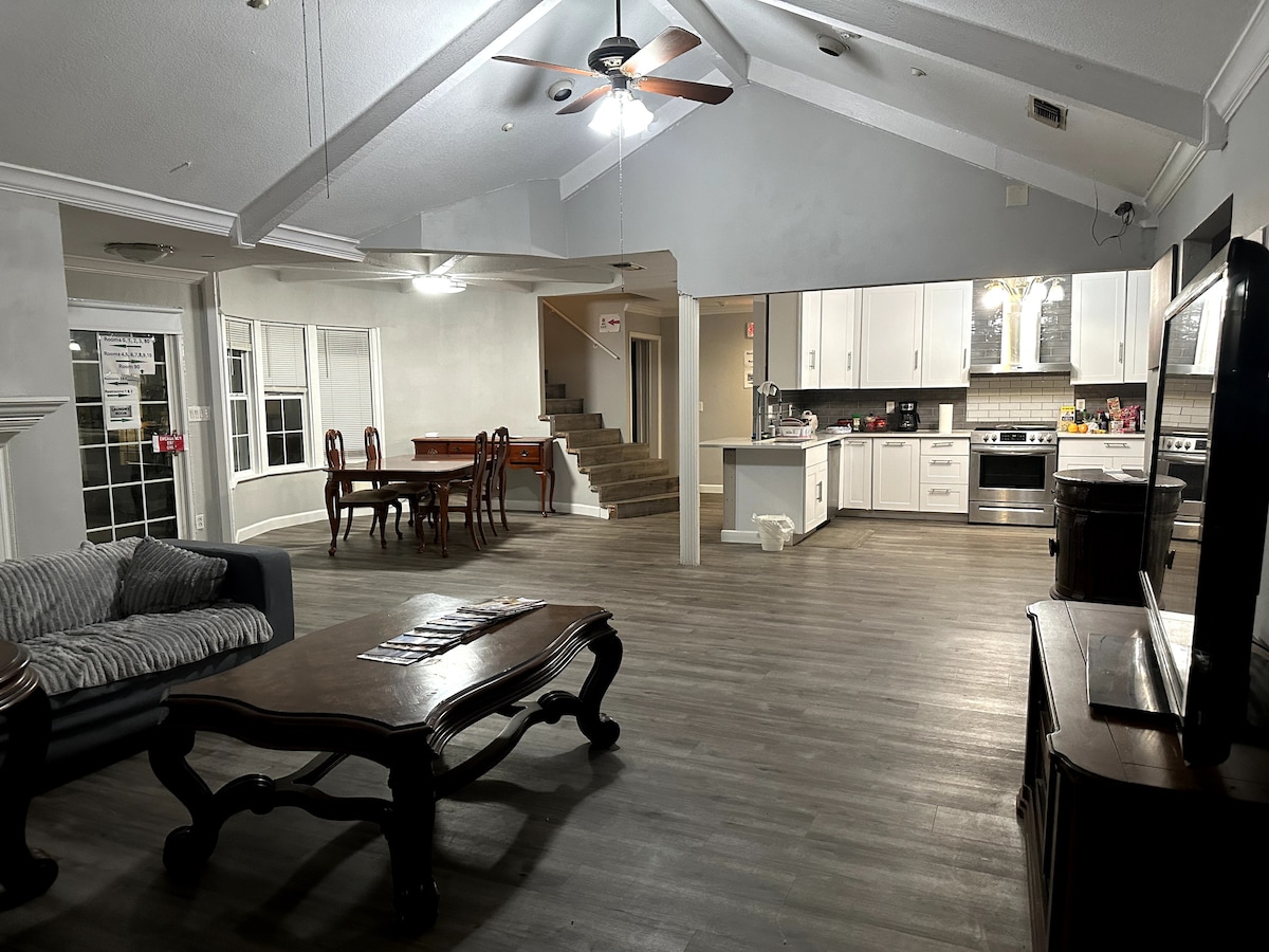 A spacious living area features high ceilings and a wooden dining table surrounded by chairs. The kitchen is visible, showcasing modern appliances and ample counter space. A comfortable gray sofa is positioned nearby, while large windows allow for natural light throughout the space.