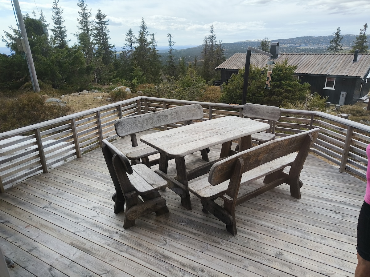 An outdoor dining area is situated on a wooden deck, featuring a robust table surrounded by four rustic benches. The surrounding natural landscape includes trees and distant hills, offering an expansive view.