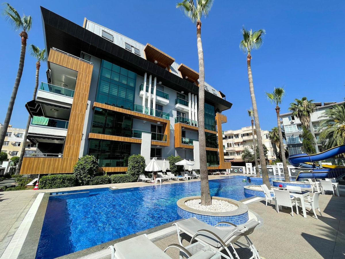 A modern apartment building features large glass windows and sleek wooden accents. A spacious swimming pool is surrounded by palm trees and comfortable sun loungers. Sun umbrellas provide shade, with a waterslide visible in the background, creating an appealing outdoor recreational area.